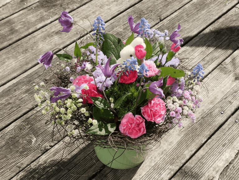 Composition de Pâques, Lapin lové dans un nid en bois naturel et tapissé d'Œillets Mousse, fleurs aux couleurs contrastées, Gypsophiles panachées, Muscaris bleus, œillets rose vif, Clématites violettes